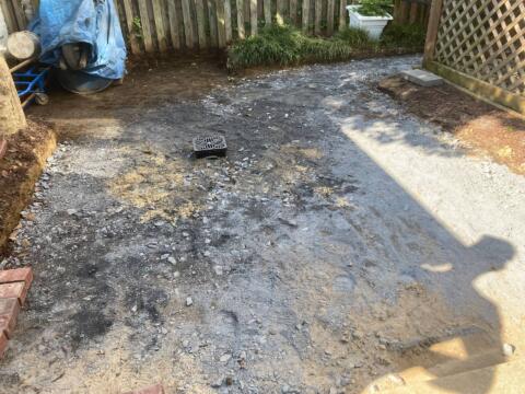 Uneven gravel yard with construction debris and drainage grate, surrounded by wooden fence.