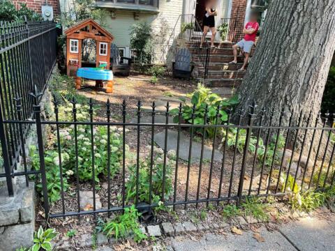 Colorful backyard with garden plants, small playhouse, and children on brick steps, surrounded by mature trees and black metal fence.