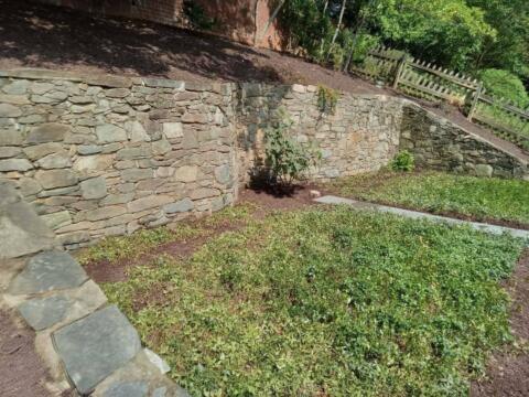 Weathered stone retaining wall in a landscaped yard with greenery and a wooden fence in the background.