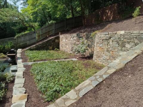 Lush landscaped backyard with stone retaining walls and green shrubbery, sunny and well-maintained.