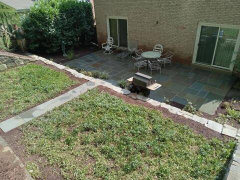 Lush backyard patio with outdoor seating and stone walkway, surrounded by greenery.