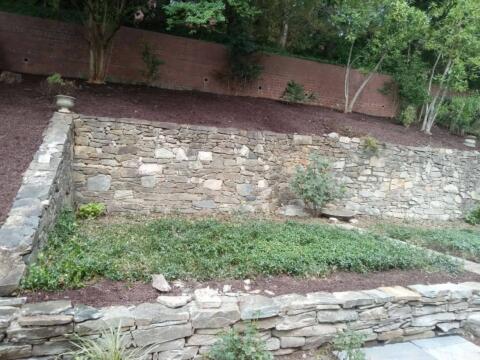 Stacked stone retaining wall with landscaped garden and trees in the background.