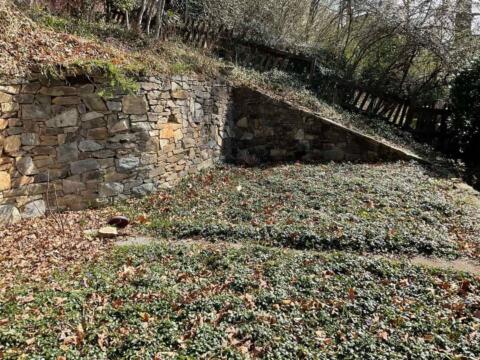 Stone retaining wall with landscaped garden below in a yard, with trees and fencing in the background.