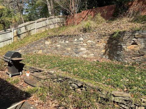 Stone retaining wall in a backyard garden with trees and fencing.