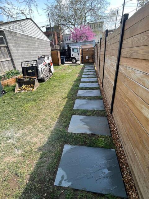 Colorful outdoor backyard with a new wooden fence and gray paver walkway, surrounded by tools and landscaping equipment.