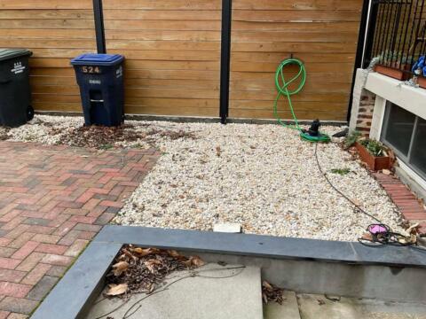 Bright green garden hose coiled on a hook in a backyard with gravel, brick, and wooden fencing.
