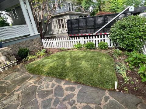 Lush green backyard lawn with stone patio and decorative garden beds, enclosed by a white picket fence.