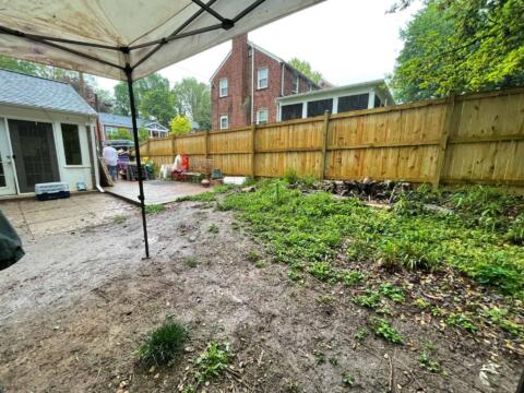 Bare backyard with dirt, fence, and garden plants under a rain shower, with a patio and houses in the background.